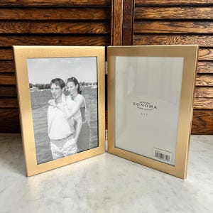 May include: Two gold-colored hinged picture frames, one open and one closed, with a black and white photo inside the closed frame. The open frame has a white mat with the text "SONOMA HOME GOODS" and is sized 5x7 inches.