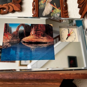 May include: A mirrored clock with a picture of a natural stone arch reflected in a body of water. The clock face has black hands and a red second hand.