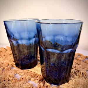 May include: Two blue drinking glasses with a faceted design. The glasses are made of a transparent material and are sitting on a woven, tan placemat. The glasses have a dark blue hue at the bottom, fading to a lighter blue at the top.