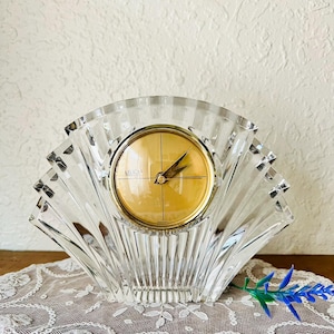 May include: A clear, fan-shaped crystal clock with a gold-colored clock face. The clock face has black hands and the word "MIKASA" is printed on the face. The clock sits on a white lace doily.