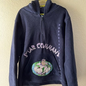 May include: A navy blue fleece hoodie with a half-zip closure and a hood. The hoodie features a large embroidered patch on the front depicting Buzz Lightyear from the Toy Story franchise. The patch is surrounded by the text "STAR COMMAND" in purple and white embroidery.