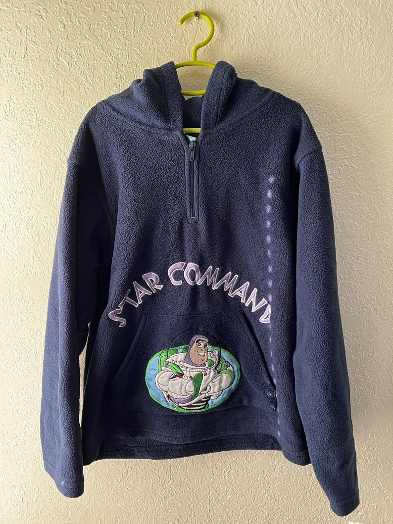 May include: A navy blue fleece hoodie with a half-zip closure and a hood. The hoodie features a large embroidered patch on the front depicting Buzz Lightyear from the Toy Story franchise. The patch is surrounded by the text "STAR COMMAND" in purple and white embroidery.