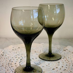 May include: Two olive-green wine glasses with rounded bowls and long stems. The base and lower portion of the bowl have a darker, textured appearance. The glasses are set on a white, lacy surface.