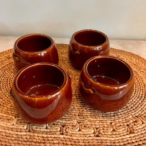 May include: A set of four small, round, brown ceramic bowls with a glossy finish. Each bowl features a smooth, curved body and two small, integrated side handles. The bowls are arranged in a square formation, showcasing their deep, uniform color.