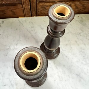 May include: Two wooden candle holders with a dark brown finish and a cream colored interior. The candle holders are on a white marble surface.