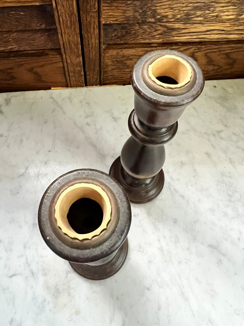 May include: Two wooden candle holders with a dark brown finish and a cream colored interior. The candle holders are on a white marble surface.