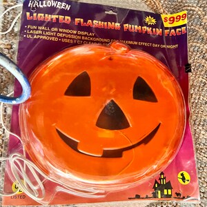 May include: A lighted flashing pumpkin face Halloween decoration. The pumpkin is orange with a black face and a white cord. The packaging says "Lighted Flashing Pumpkin Face" and "UL Approved".