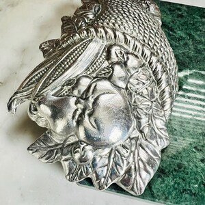 May include: Silver serving dish shaped like a cornucopia overflowing with fruit and vegetables. The dish is on a green marble surface.