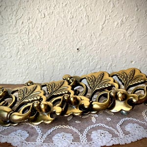 May include: Ornate, antique brass drawer pull with a floral and leaf design. The hardware is set against a white lace doily and a textured wall, showcasing its vintage aesthetic. The pull is approximately 10 inches long.