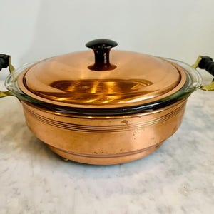 May include: A copper cooking pot with a glass lid and black handle. The pot has a round shape with a copper finish and brass handles. The lid has a black knob. The pot is designed for cooking.