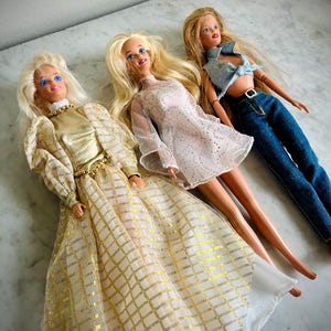 May include: Three vintage fashion dolls with blonde hair and blue eyes. One doll wears a gold and white gown, another a pink dress, and the third a denim top and jeans. The dolls are lying down on a light-colored surface.