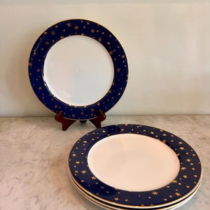 May include: A set of round ceramic dinner plates featuring a solid white center and a wide navy blue rim decorated with a pattern of small gold stars. The edges of the plates are finished with a thin gold trim, providing a consistent design.