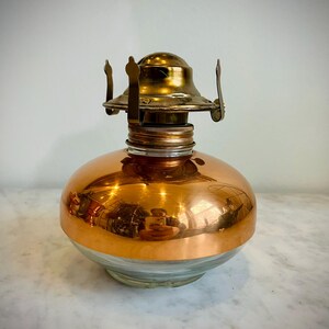 May include: A vintage oil lamp with a clear glass base and a copper-colored metal top and body. The lamp features a brass burner assembly and a reflective surface, creating a warm, antique aesthetic. The lamp is designed to hold oil for illumination.