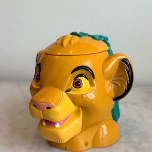 May include: A yellow Simba-shaped container with a lid. The container has a pink nose, white teeth, and a green mane. The eyes are brown and yellow. The container is made of plastic and is likely a collectible item.
