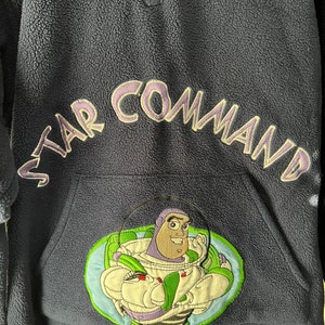 May include: A dark blue fleece pullover with a zip fastening. The front of the pullover features an embroidered image of Buzz Lightyear from the Toy Story franchise. The text "STAR COMMAND" is embroidered in purple on the right shoulder.