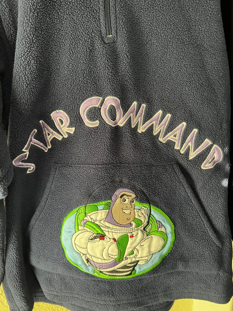 May include: A dark blue fleece pullover with a zip fastening. The front of the pullover features an embroidered image of Buzz Lightyear from the Toy Story franchise. The text "STAR COMMAND" is embroidered in purple on the right shoulder.