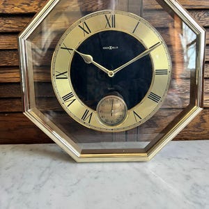 May include: A gold-toned octagonal wall clock with a black face and Roman numeral hour markers. The clock has a clear glass cover and a smaller, round, silver-toned dial with black markings for seconds.