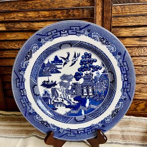 May include: A blue and white Willow pattern plate. The plate features a detailed landscape scene with pagodas, bridges, and birds. The plate is displayed on a wooden stand, showcasing its intricate design and classic aesthetic.