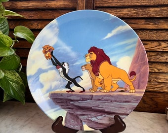 Może przedstawiać: Ceramiczny talerz przedstawiający scenę z filmu Disneya Król Lew. Talerz przedstawia Simbę, Mufasę i Rafikiego na klifie z widokiem na sawannę. Talerz jest niebieski i biały z brązowym drewnianym stojakiem.