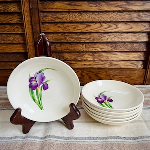 May include: Set of six white ceramic bowls with a purple iris flower design. The bowls are stacked on top of each other and are on a striped fabric surface.