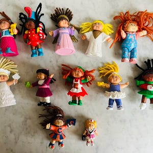 May include: A collection of vintage Cabbage Patch Kids dolls, each with unique hairstyles and outfits. The dolls are in various poses, wearing dresses, overalls, and other colorful attire. Some dolls are holding accessories.