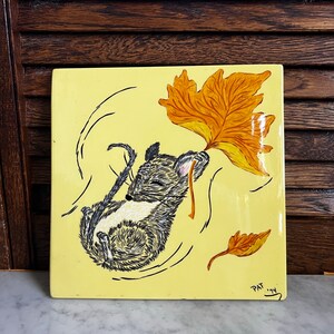 May include: A yellow ceramic tile with a hand-painted illustration of a gray and white mouse holding a brown leaf. The mouse is surrounded by swirling lines and two additional brown leaves. The tile is signed "PAT '74" in the bottom right corner.
