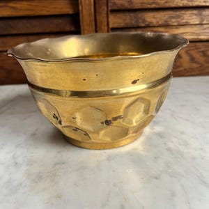 May include: A small, antique brass bowl with a hammered texture and a decorative band. The bowl has a scalloped rim and a warm, golden color. It is likely used for decorative purposes.