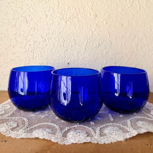 May include: Three cobalt blue stemless wine glasses. The glasses are round with a textured surface and sit on a white lace doily. The glasses are empty and reflect the light.