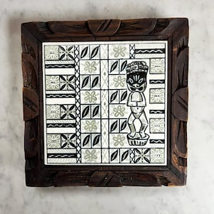 May include: A wooden framed tile with a black and white geometric design and a green and white tiki figure in the center.
