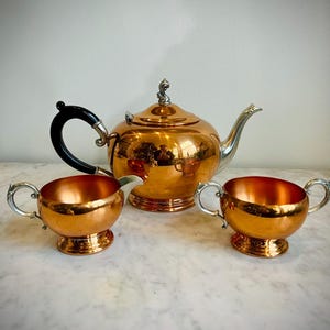 May include: A copper tea set with a teapot and two matching cups. The teapot has a black handle and a silver spout. The cups have silver handles. The set is displayed on a marble surface.