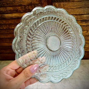 May include: A clear glass plate with a scalloped edge and a detailed sunburst pattern. The plate has a central circular indentation and is decorated with a delicate leaf design around the rim. The plate is held by a person.
