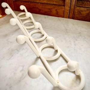 May include: Off-white plastic wall-mounted coat rack with a unique design. The rack features a series of curved hooks and circular elements, creating a decorative and functional storage solution. The rack is designed to hold multiple items.