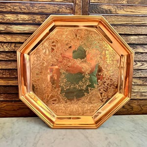 May include: An octagonal copper-colored tray with ornate floral engravings. The tray has a raised edge and a polished finish, reflecting the surrounding environment. The tray is likely used for serving or display purposes.