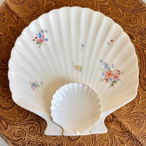 May include: A white ceramic dish shaped like a large scallop shell, featuring delicate floral patterns in pink, blue, and yellow. A smaller, matching shell-shaped piece is attached to the front, accented with a thin gold rim along its edge.