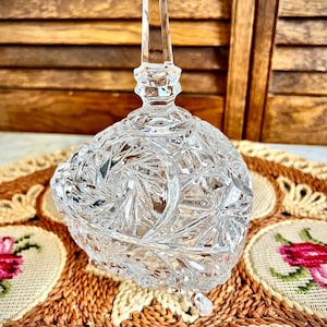 May include: A clear glass trinket dish with a cut crystal design and a pointed lid. The dish has three small feet and is sitting on a woven placemat.