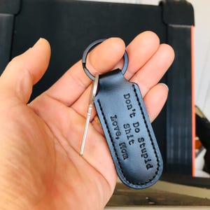 May include: Black leather keychain with a metal key and a black metal ring. The keychain has the text "Don't Do Stupid Shit Love, Mom" embossed on it. The keychain is held in a hand.