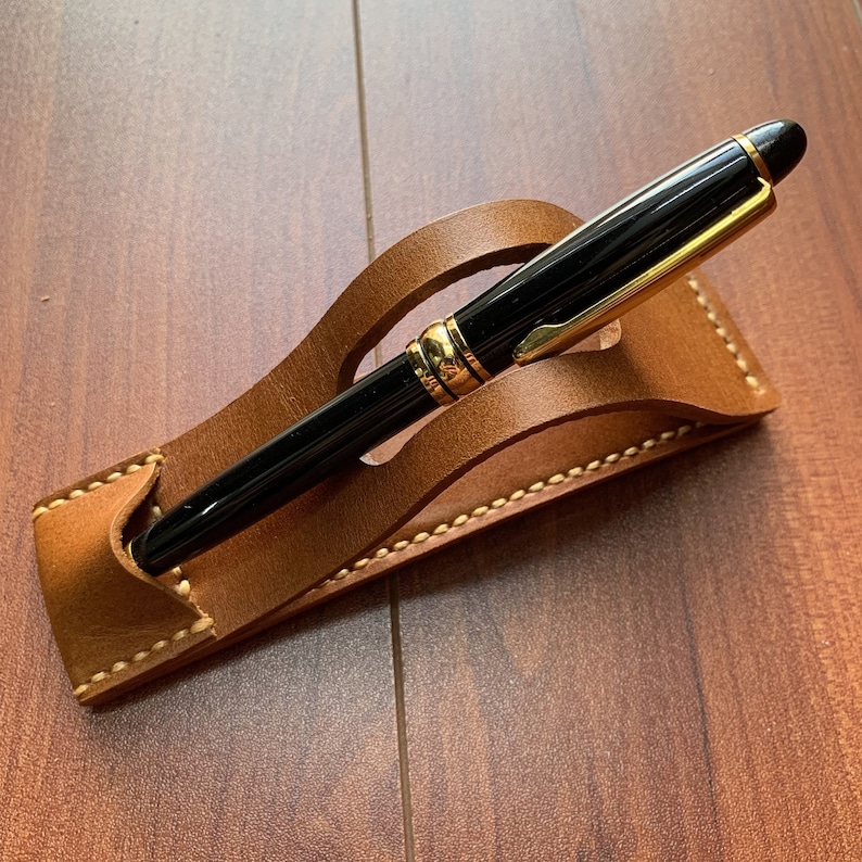 May include: A brown leather pen holder with a black pen resting inside. The pen holder has a decorative stitching detail.