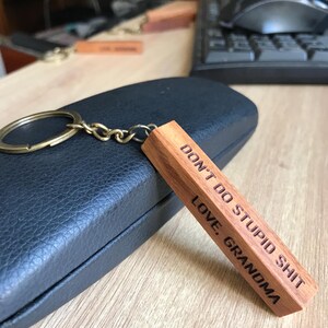May include: A wooden keychain with a brass ring and chain. The rectangular wooden piece is engraved with the words "DON'T DO STUPID SHIT" and "LOVE, GRANDMA" in a bold, sans-serif font. The keychain rests on a black leather case.