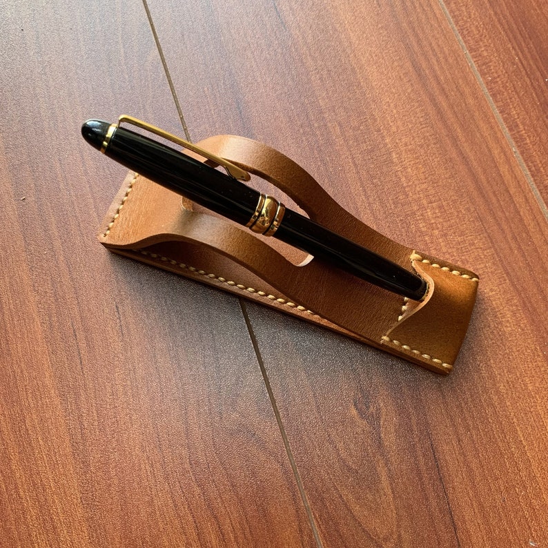 May include: A brown leather pen holder with a stitched design. A black pen with gold accents is resting in the holder.