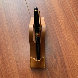 May include: A brown leather pen holder with a black pen inside. The pen holder has a stitched design and a curved shape.