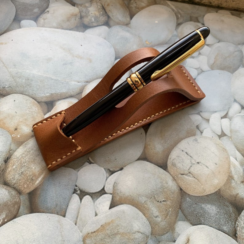 May include: A brown leather pen holder with a stitched edge. A black pen with gold accents is resting in the holder.