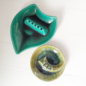 May include: Two vintage ceramic ashtrays. One is green and shaped like a leaf with a rectangular depression for cigarettes. The other is green and brown with a fish design.