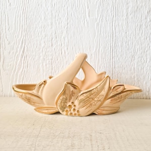 May include: A ceramic dish shaped like a flower with a gold handle and gold leaf accents. The dish is a pale pink color.