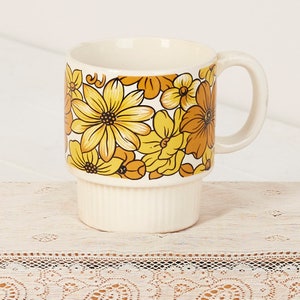 Op de afbeelding: Een witte keramische mok met een gestapeld ontwerp. De mok heeft een bloemenpatroon met gele, oranje en bruine bloemen op een witte achtergrond.
