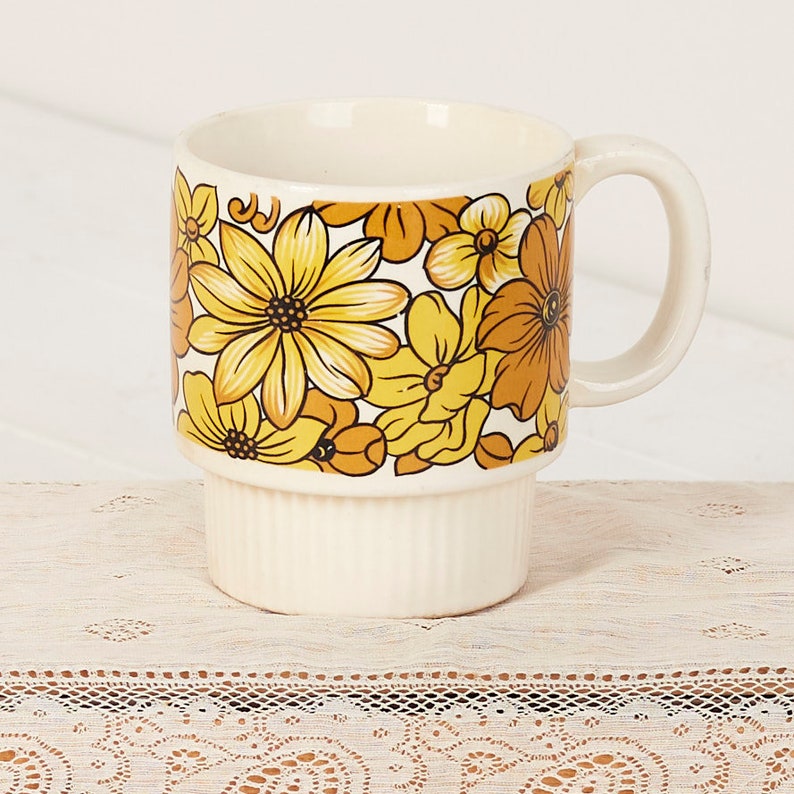 Op de afbeelding: Een witte keramische mok met een gestapeld ontwerp. De mok heeft een bloemenpatroon met gele, oranje en bruine bloemen op een witte achtergrond.