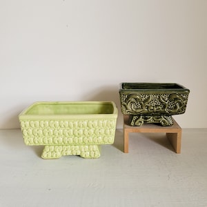 May include: Two ceramic planter pots with raised designs. The larger pot is light green with a textured pattern and four legs. The smaller pot is dark green with a floral design and four legs. Both pots are on a wooden stand.