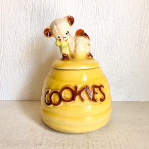 May include: A vintage yellow ceramic cookie jar with a lid featuring a beige bear figurine. The jar has a beehive design with the word "COOKIES" in brown lettering. The bear has brown ears and a green scarf.