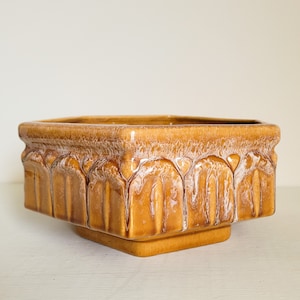 May include: A square ceramic planter with a brown glaze and white accents. The planter has a geometric design with raised edges.