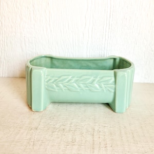 May include: A rectangular, light green ceramic planter with a raised leaf design. The planter has a ribbed design on the sides and is elevated by two short feet. The interior is a matching light green color.