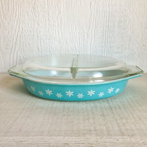 May include: A turquoise Pyrex casserole dish with a clear glass lid. The dish features a snowflake pattern in white around the rim. The lid has a handle and a divider. A vintage kitchenware item.